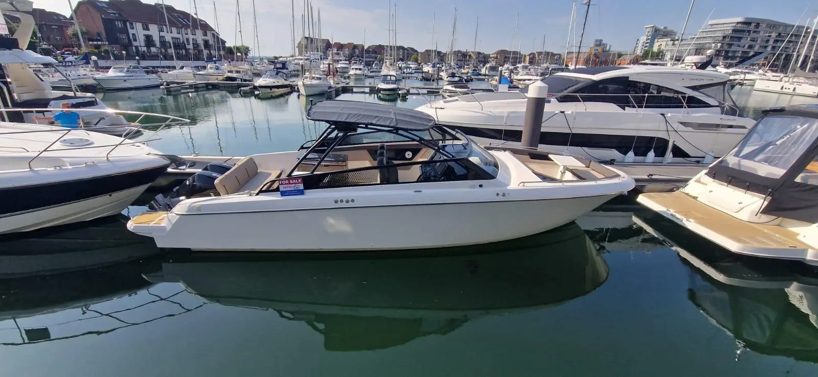 A white boat marked "FOR SALE" is docked among several other boats in a marina with calm waters and sunny weather.